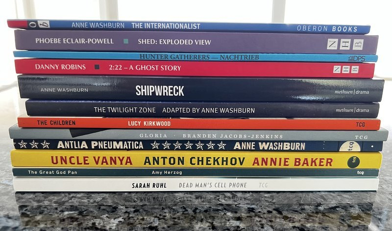 A tall stack of play scripts on the kitchen counter, including The Internationalist, Shipwreck, The Children, Gloria, Uncle Vanya, and Dead Man's Cell Phone