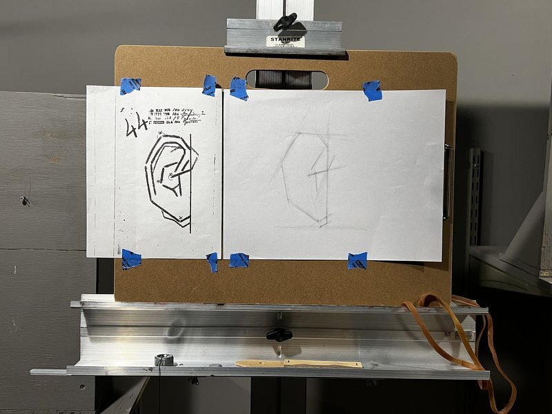 A drawing board on my easel with a printed reference diagram of an ear taped beside my faint pencil block-in of the same ear