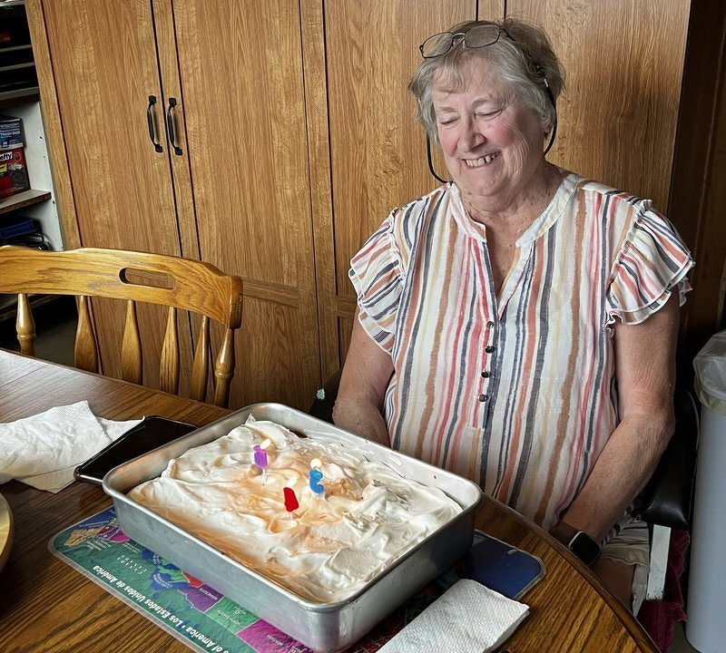 My mom smiling at the kitchen table behind a pan of frosted birthday cake with candles