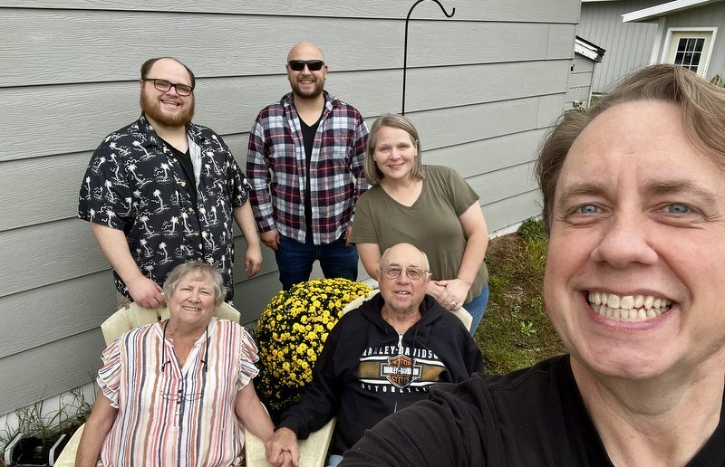 A family selfie outside my parents' house, with my mom and dad seated beside a big pot of yellow mums and the rest of us gathered around them
