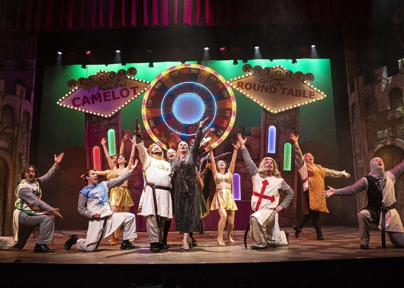 The Spamalot cast mid-number on the NorShor stage, arms flung wide in front of glowing Welcome to Fabulous Camelot and Knights of the Round Table signs and a giant roulette wheel