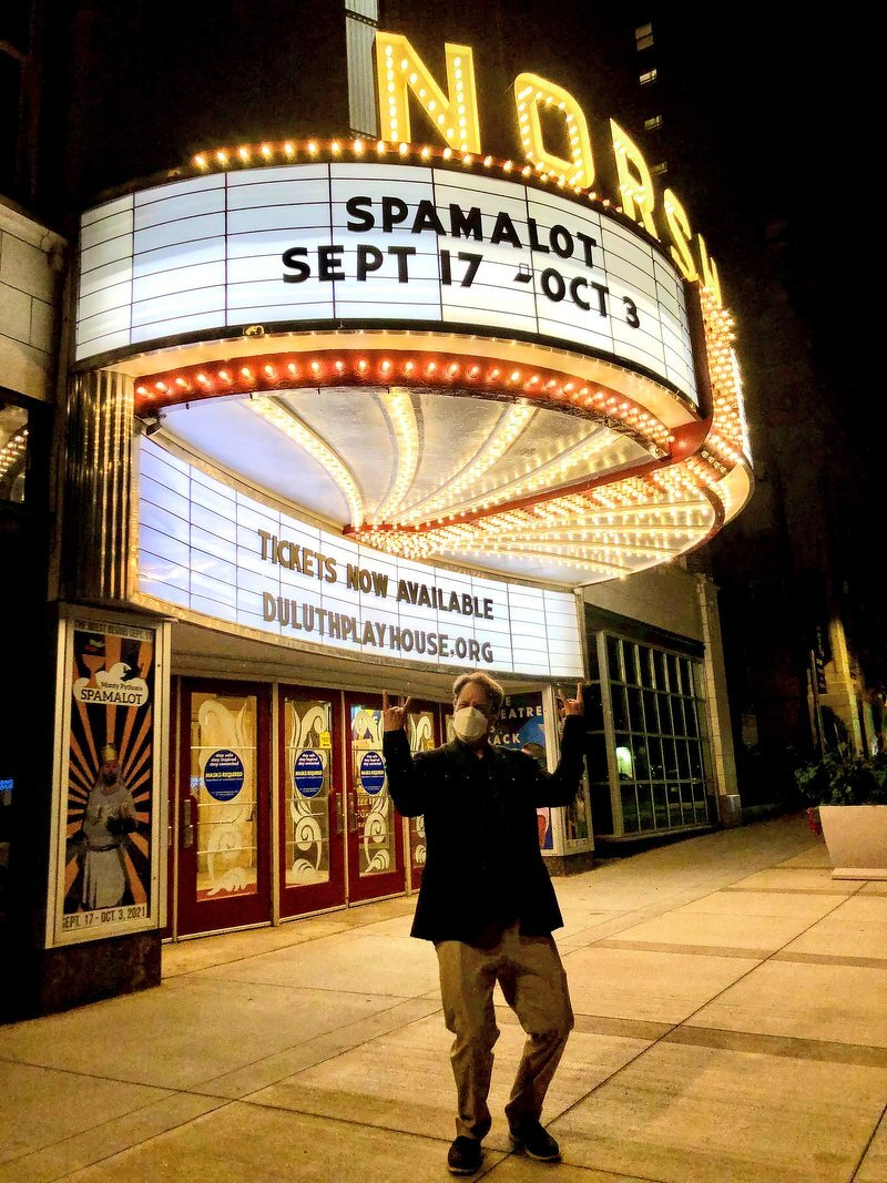 Justin, masked, throwing up rock horns under the glowing NorShor Theatre marquee reading Spamalot, Sept 17 - Oct 3