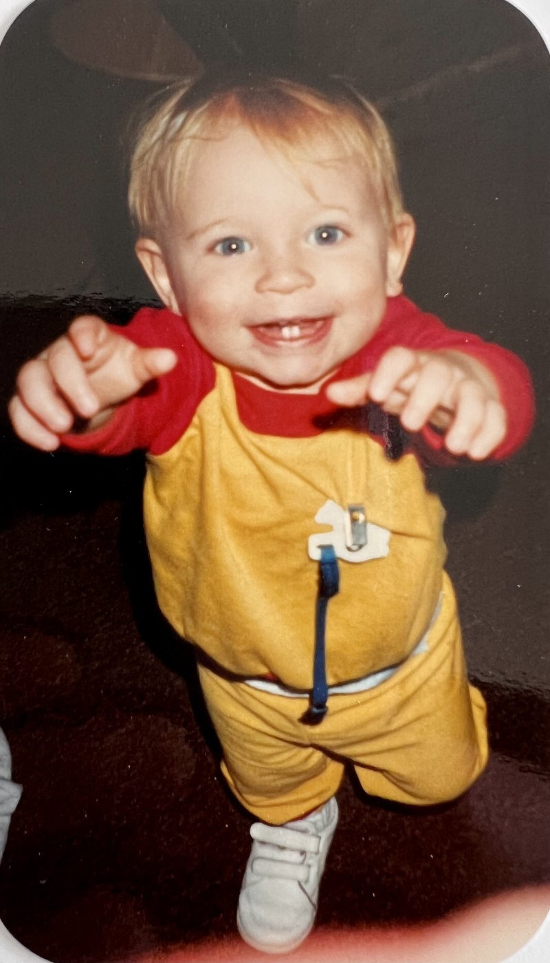 Baby Alex in a yellow romper, grinning and reaching toward the camera