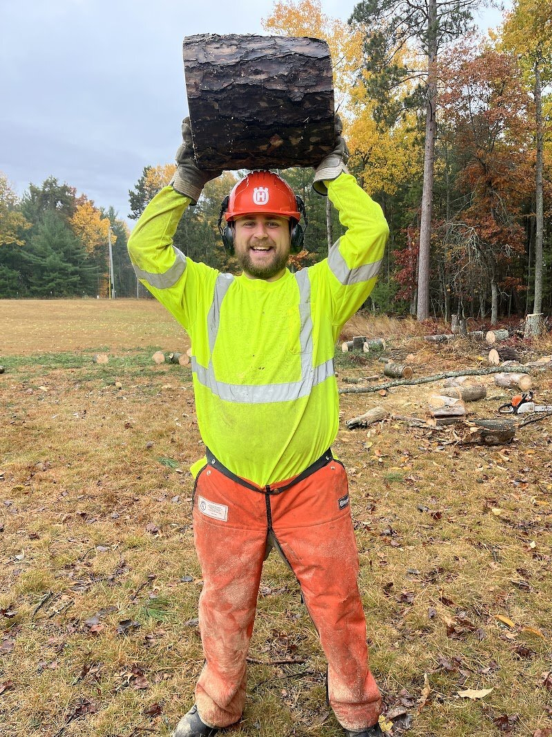 Alex in a hard hat, high-vis jacket and chainsaw chaps, hoisting a giant log over his head
