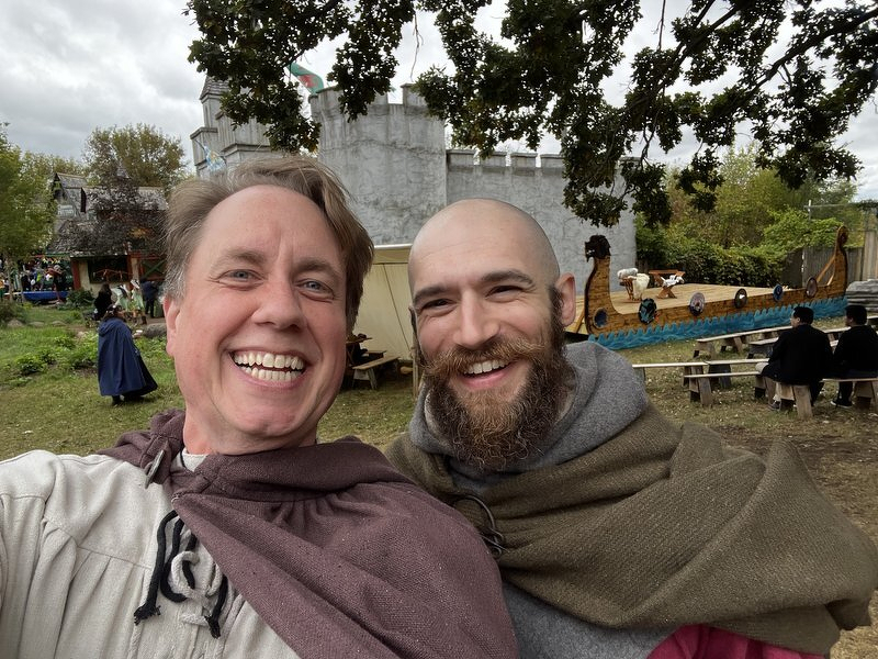Justin and Matt in wool cloaks, laughing, with a longship and picnic tables behind them