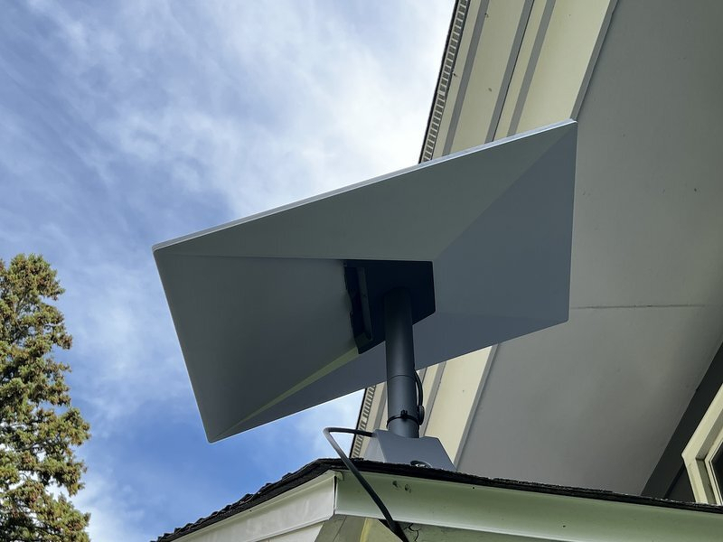 The Starlink dish mounted on the side of the house, tipped up toward a blue sky