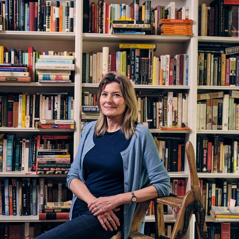 Jennifer Egan seated on a wooden ladder in front of floor-to-ceiling bookshelves