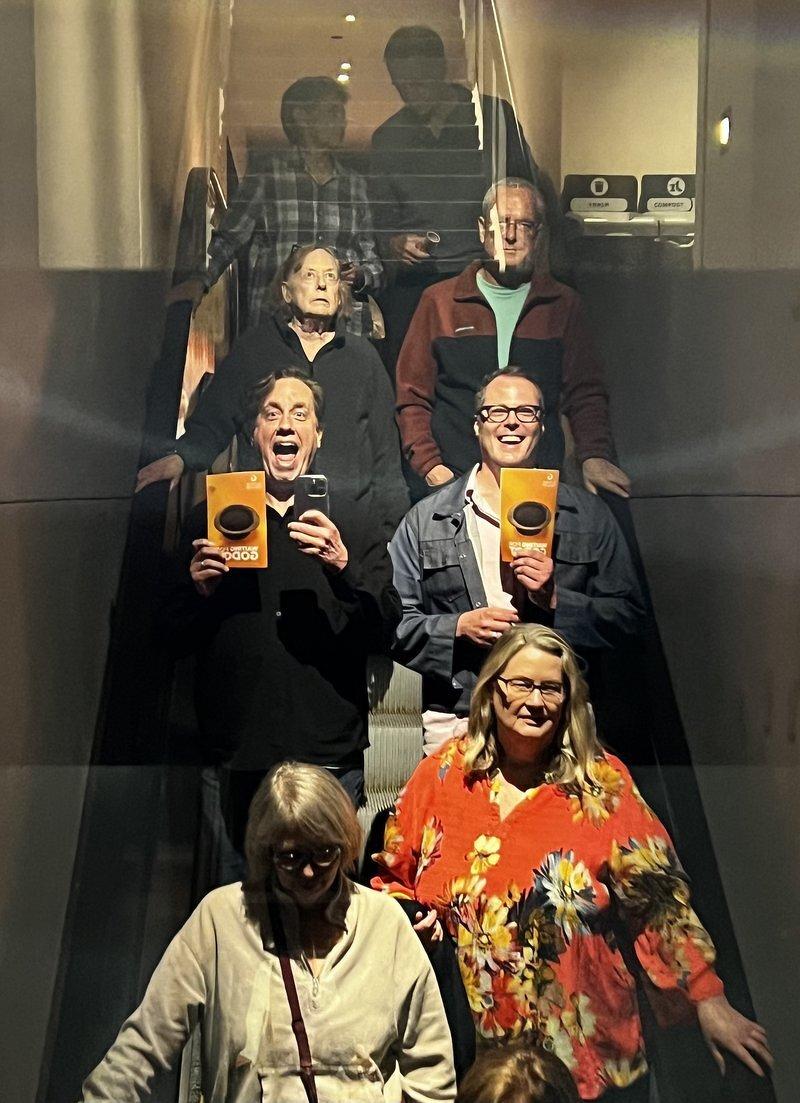 A mirrored-ceiling selfie of Justin and Jason riding the Guthrie escalator with their Godot programs, surrounded by other theatergoers