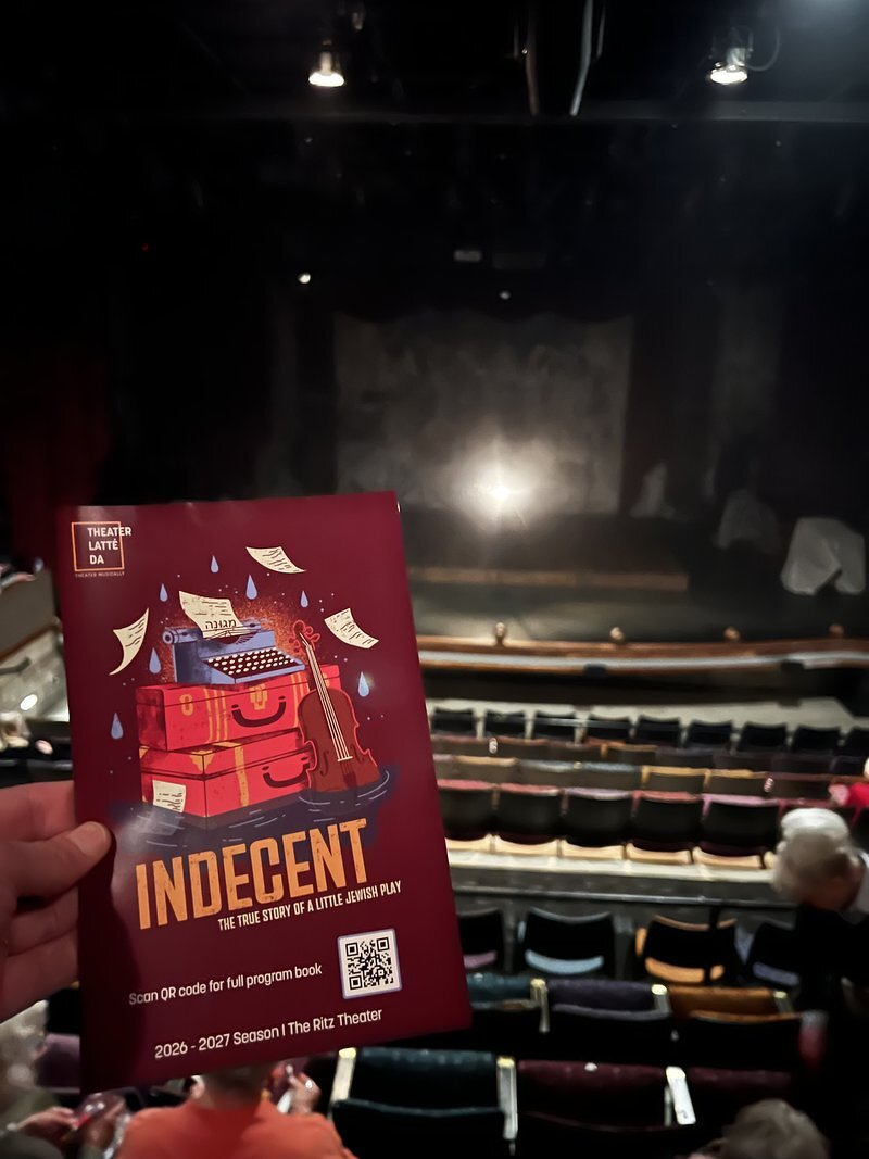 The Indecent program, with a pile of suitcases and a typewriter on the cover, held up in front of the stage at Theater Latté Da