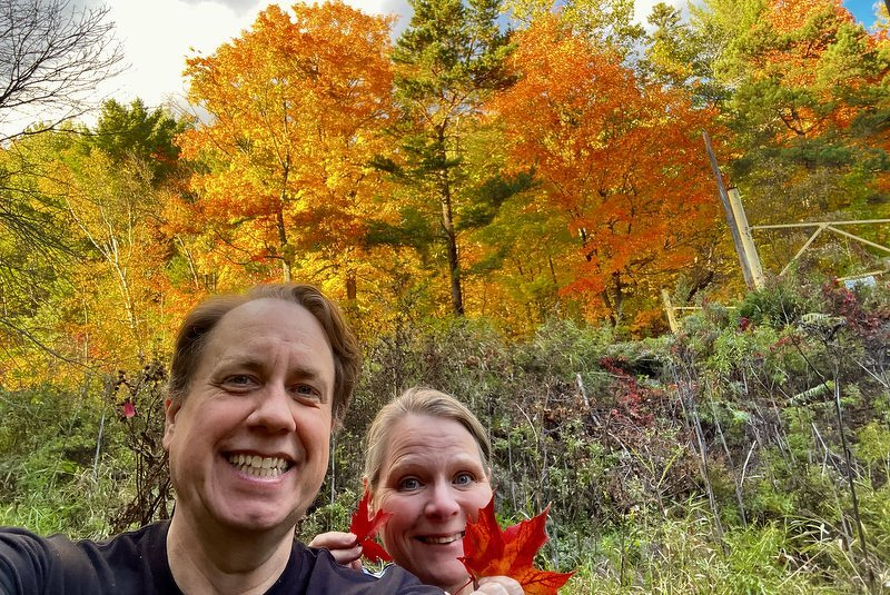 A selfie of Justin and Jody, who is holding up a bright red maple leaf, in front of a hillside of orange and gold trees