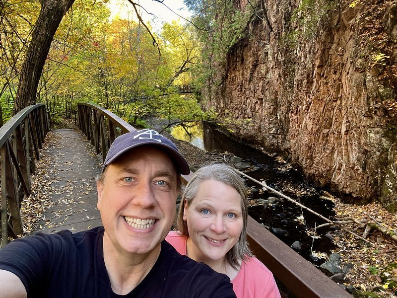 A selfie of Justin and Jody on a wooden footbridge, a towering rock wall and golden fall trees behind them