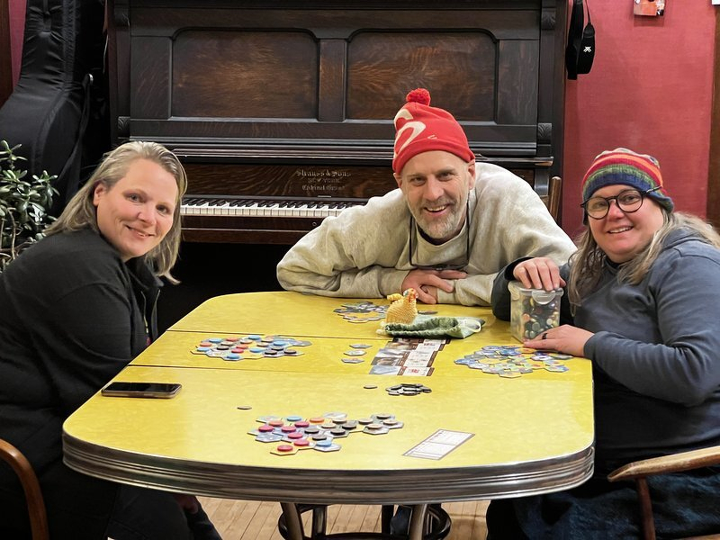 Jody, Tom, and Brandy around a table covered in Cascadia tiles, an upright piano behind them