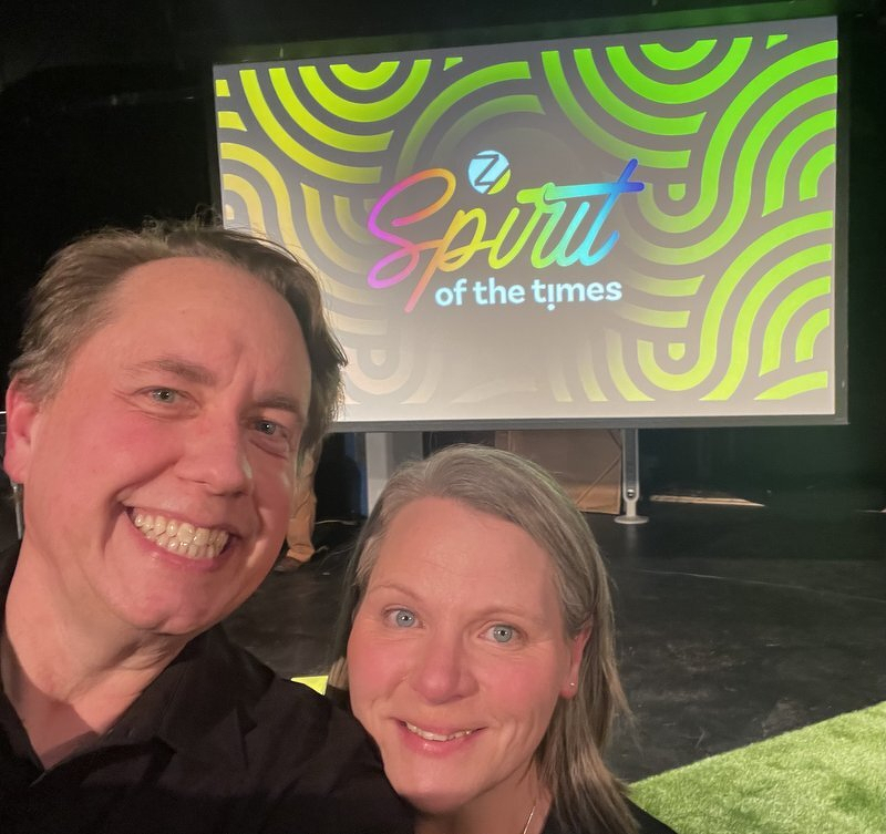 A selfie of Justin and Jody in front of a screen reading Spirit of the Times in swirling neon green and yellow