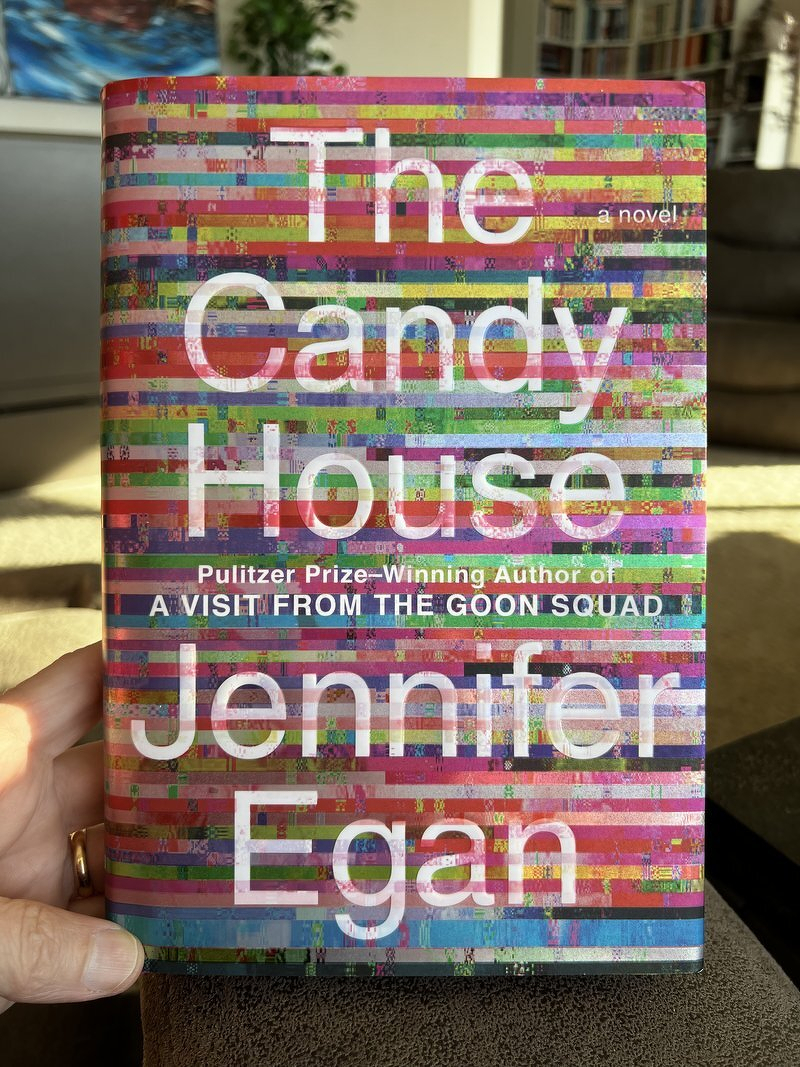 Holding up a hardcover copy of The Candy House by Jennifer Egan, its title over a glitchy rainbow of horizontal stripes