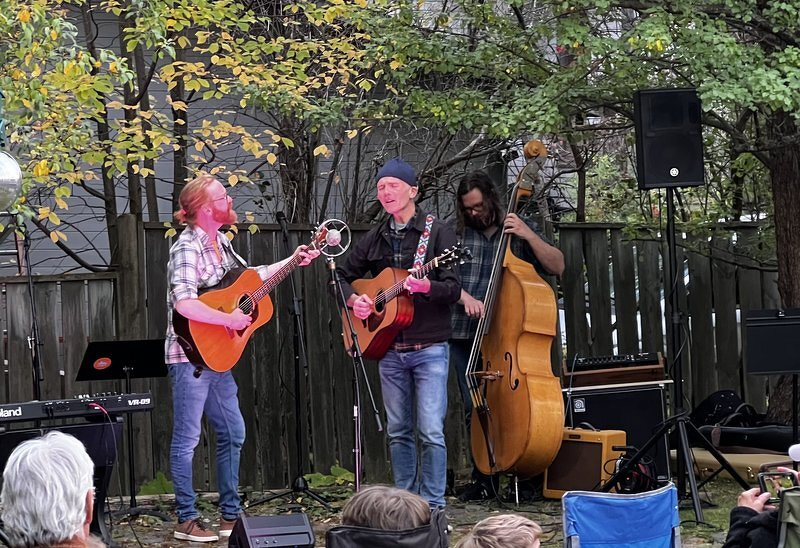 A trio with two acoustic guitars and an upright bass playing in a fenced backyard under trees, audience members in camp chairs in the foreground