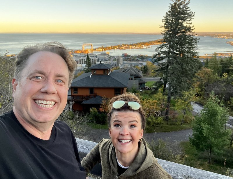 A selfie of Justin and Kate at the top of the incline, Duluth rooftops and Lake Superior glowing at sunset behind them