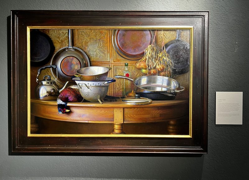 A Jeffrey Larson painting of copper pots, a colander, and kitchenware crowded on a wooden table before an ornate tin backdrop