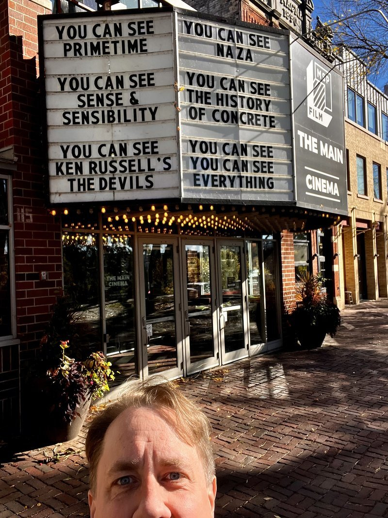 Justin peeking into the frame below the Main Cinema marquee, which jokingly reads You Can See Primetime, You Can See Sense and Sensibility, You Can See Ken Russell's The Devils, and You Can See You Can See Everything