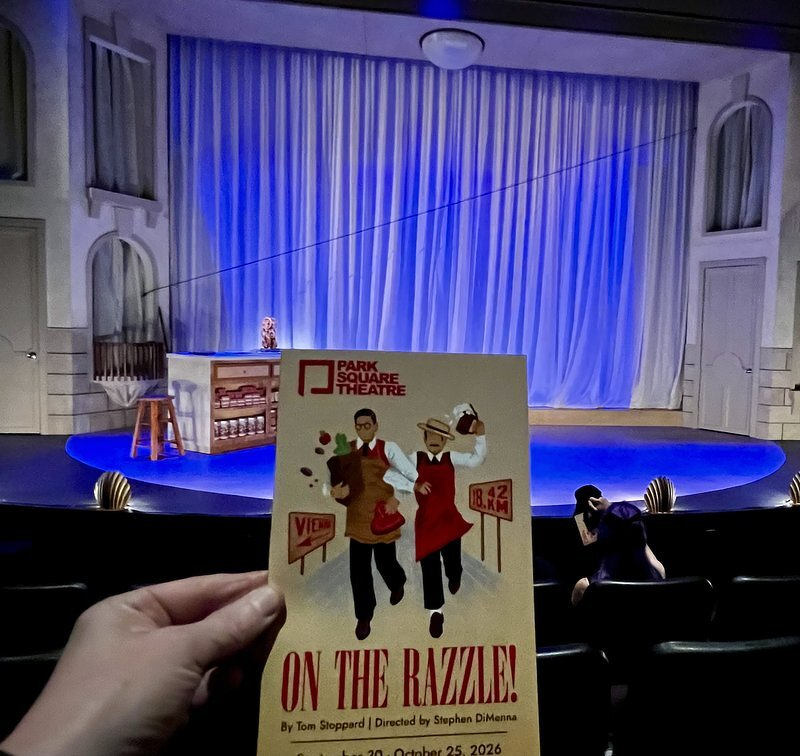 Holding up a program for On the Razzle in front of the Park Square Theatre stage, lit in blue with a rustic shop set