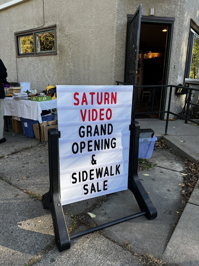A sidewalk sign reading Saturn Video Grand Opening and Sidewalk Sale outside a small stucco storefront