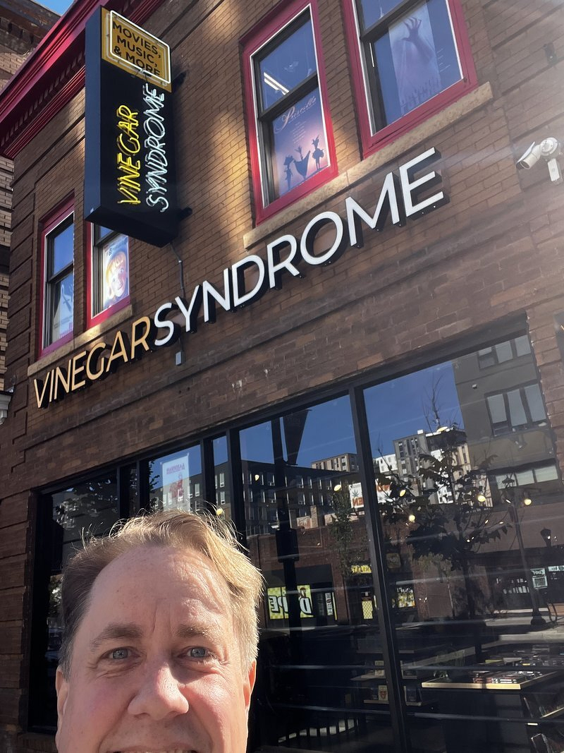 Justin peeking into the frame below the Vinegar Syndrome sign on a brick storefront in Dinkytown