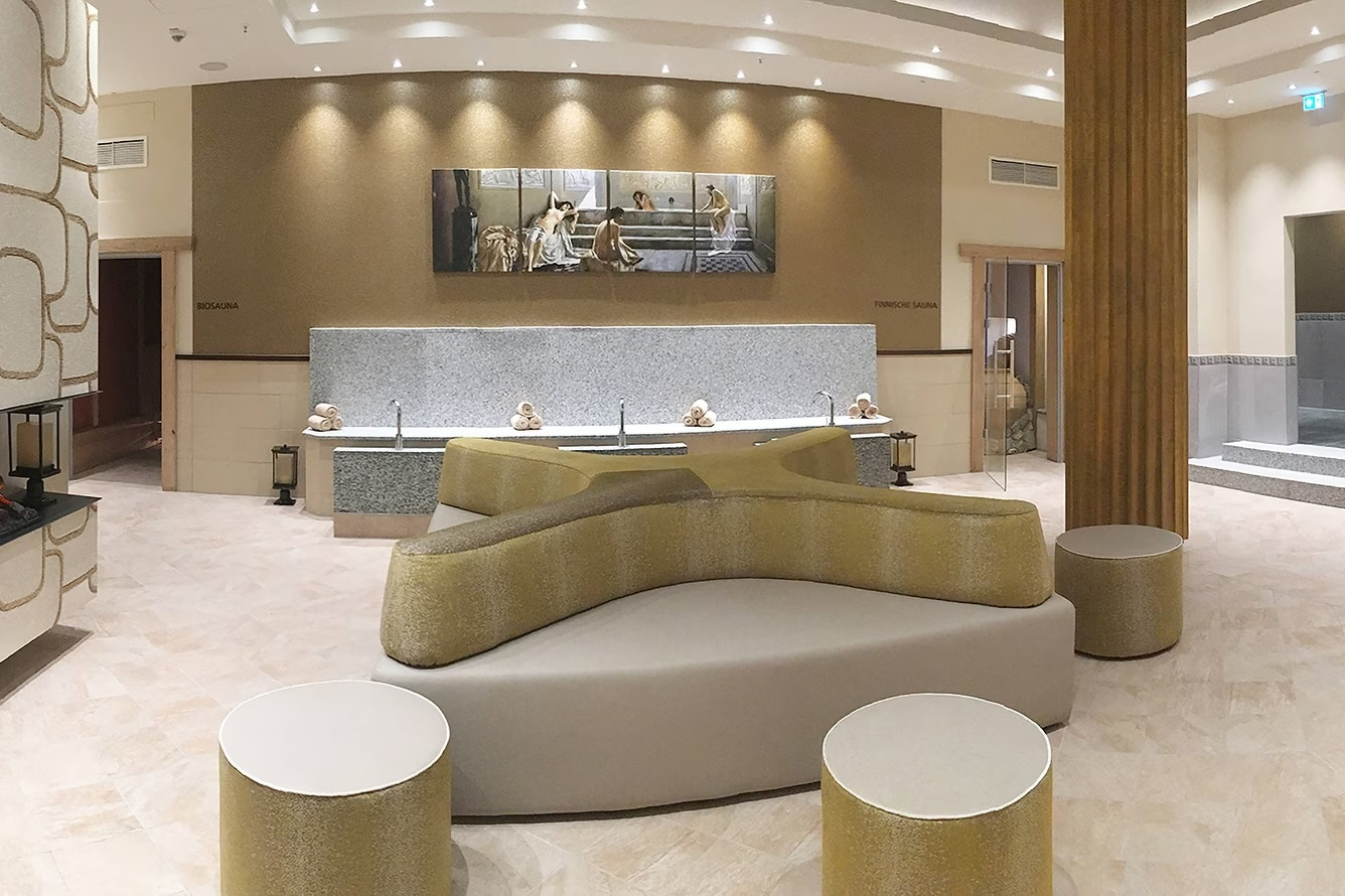 A modern, spacious wellness and relaxation area at the Visiolife in Bonn, Germany. The room features neutral beige tones, soft recessed lighting, and a large, curved grey sectional sofa in the centre. In the background, there is a long vanity with multiple sinks and a horizontal piece of artwork on the wall. The space is finished with light-coloured stone flooring and includes a fireplace on the left and a staircase leading to another section on the right.