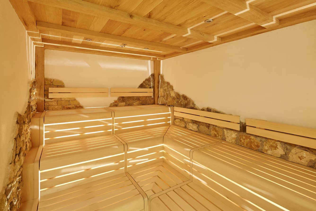 A modern Finnish sauna interior with light-coloured wooden tiered seating arranged in an L-shape. The walls feature a unique design of smooth white plaster interspersed with rough, natural stone sections that appear to break through the surface. Recessed lighting in the wooden slatted ceiling creates a warm, inviting glow, casting soft shadows across the benches.