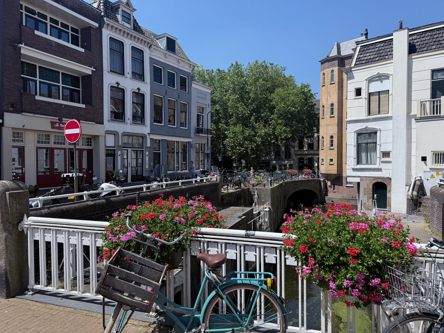 A peaceful canal scene showcases a charming row of traditional buildings, vibrant flower baskets on a white railing, and a parked bicycle under a clear blue sky.