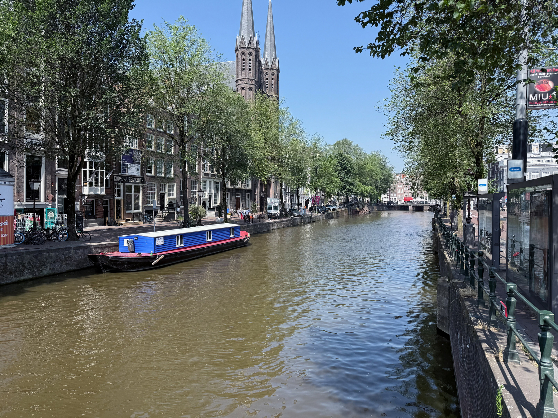 A canal lined with trees and buildings has a small boat with a blue and red cabin floating on it under a clear blue sky.