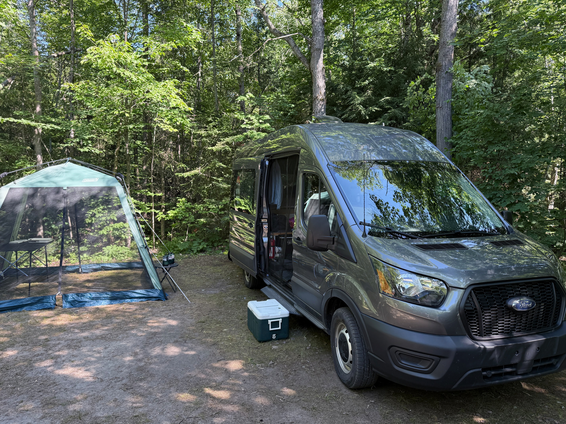 A gray Ford camper van is parked in a wooded area next to a green tent with mesh walls.