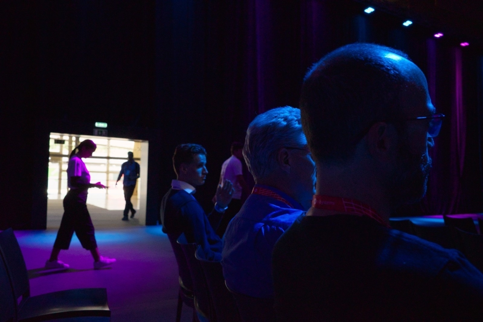 Silhouetted individuals are seated in a dimly lit room with purple lighting, while others are seen walking through a lit doorway.