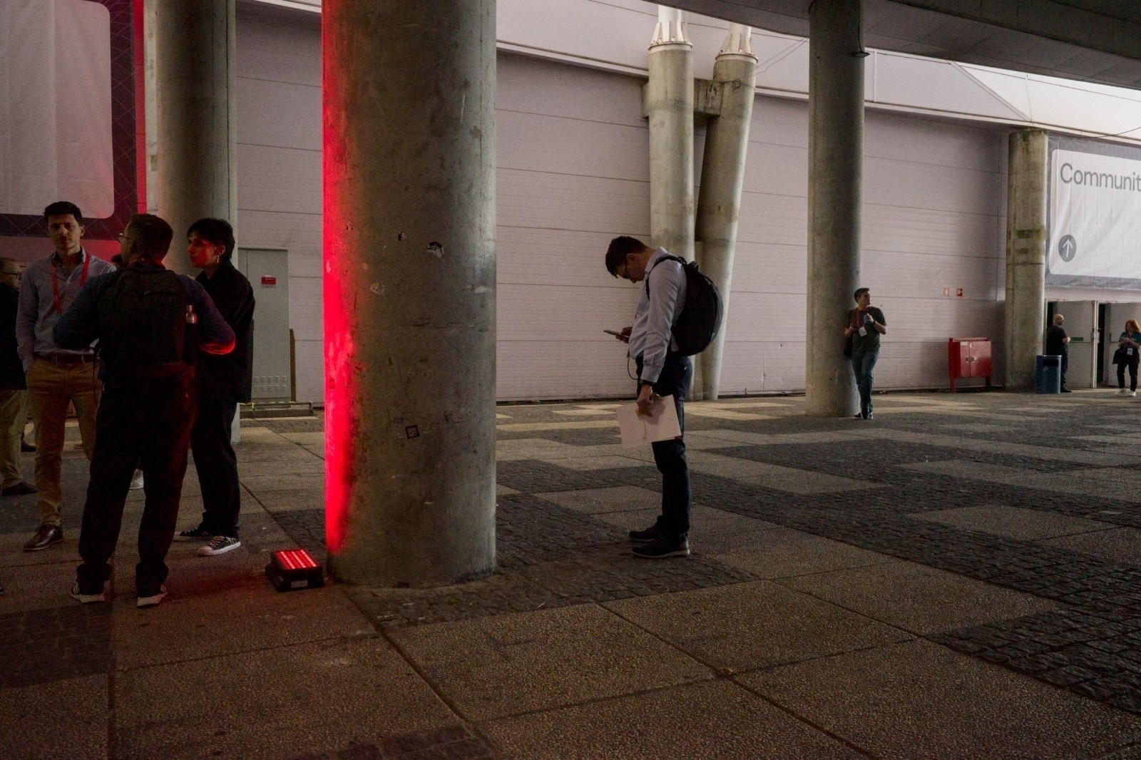 A man with a backpack is standing alone checking his phone under a red light, while a group of people interacts nearby under a covered outdoor area.