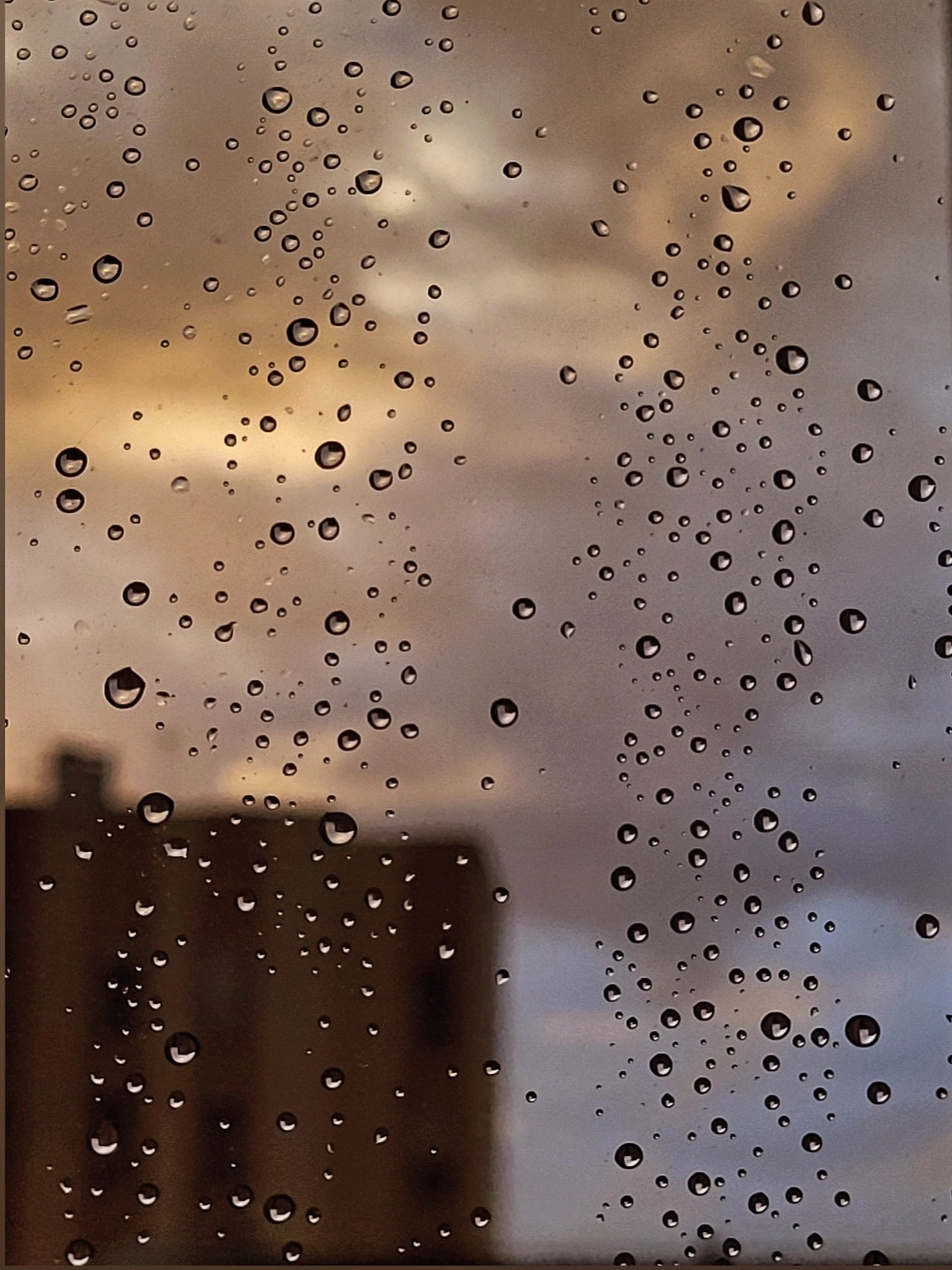 Raindrops are scattered on a window with a blurred view of a building and a cloudy, orange-tinted sky in the background.