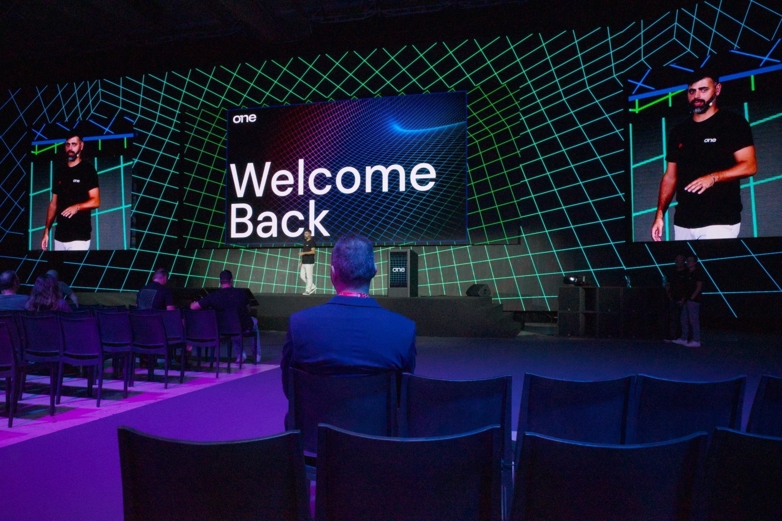 A speaker is presenting on stage with a Welcome Back message displayed on large screens to an audience in a spacious, futuristic venue.