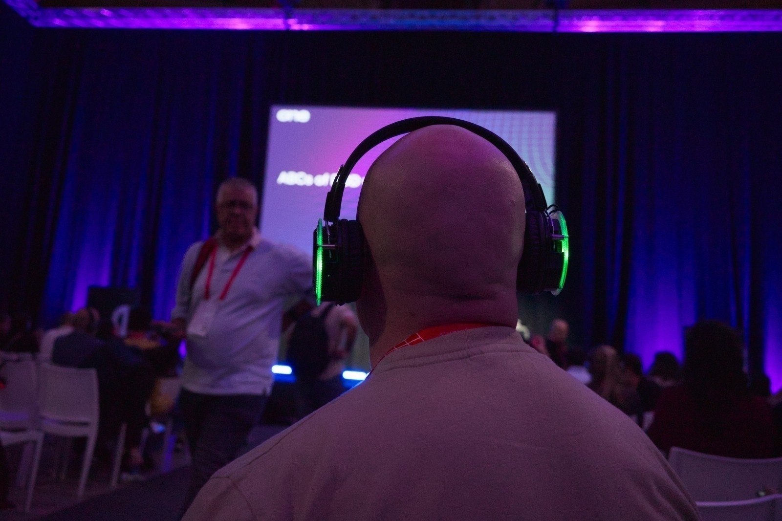 A person wearing green-lit headphones is seated in a dimly lit room with people and a screen in the background displaying text.
