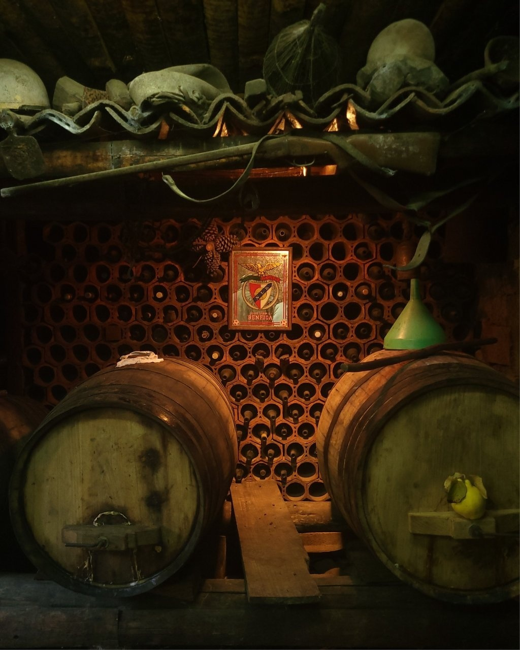 A rustic cellar features two large wooden barrels and a wall of bottle compartments, with various vintage items and a poster above.