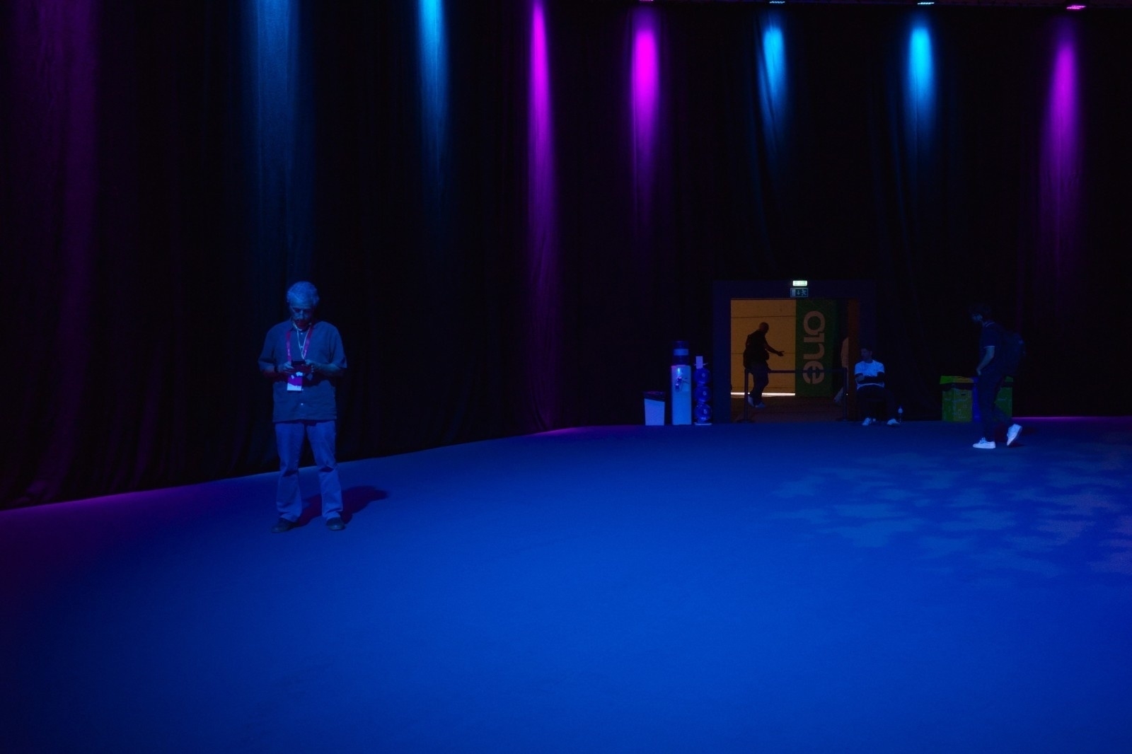 A dimly lit room with a blue floor, illuminated by colored lights and a few people visible in the background.
