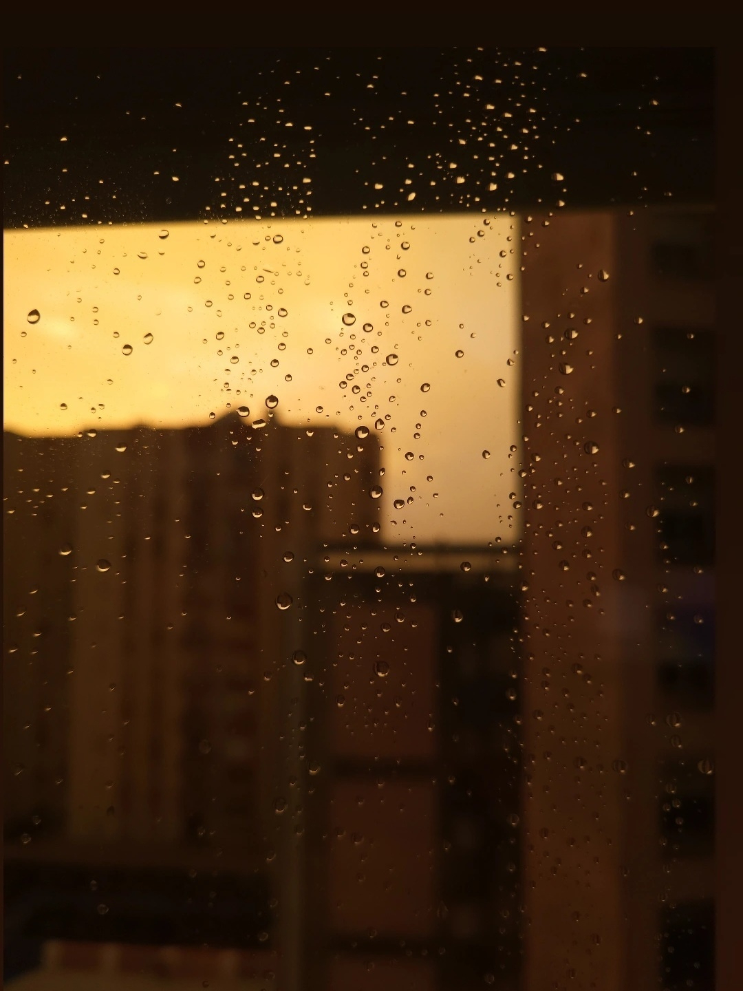 Raindrops are scattered on a window in front of a city skyline during an orange sunset.