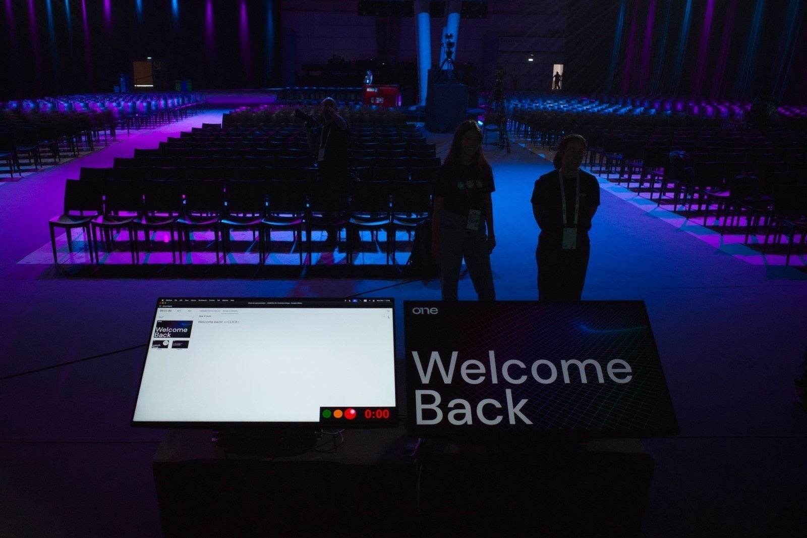 A stage with a Welcome Back message and a computer monitor is set up in a large, dimly lit auditorium with rows of empty chairs.