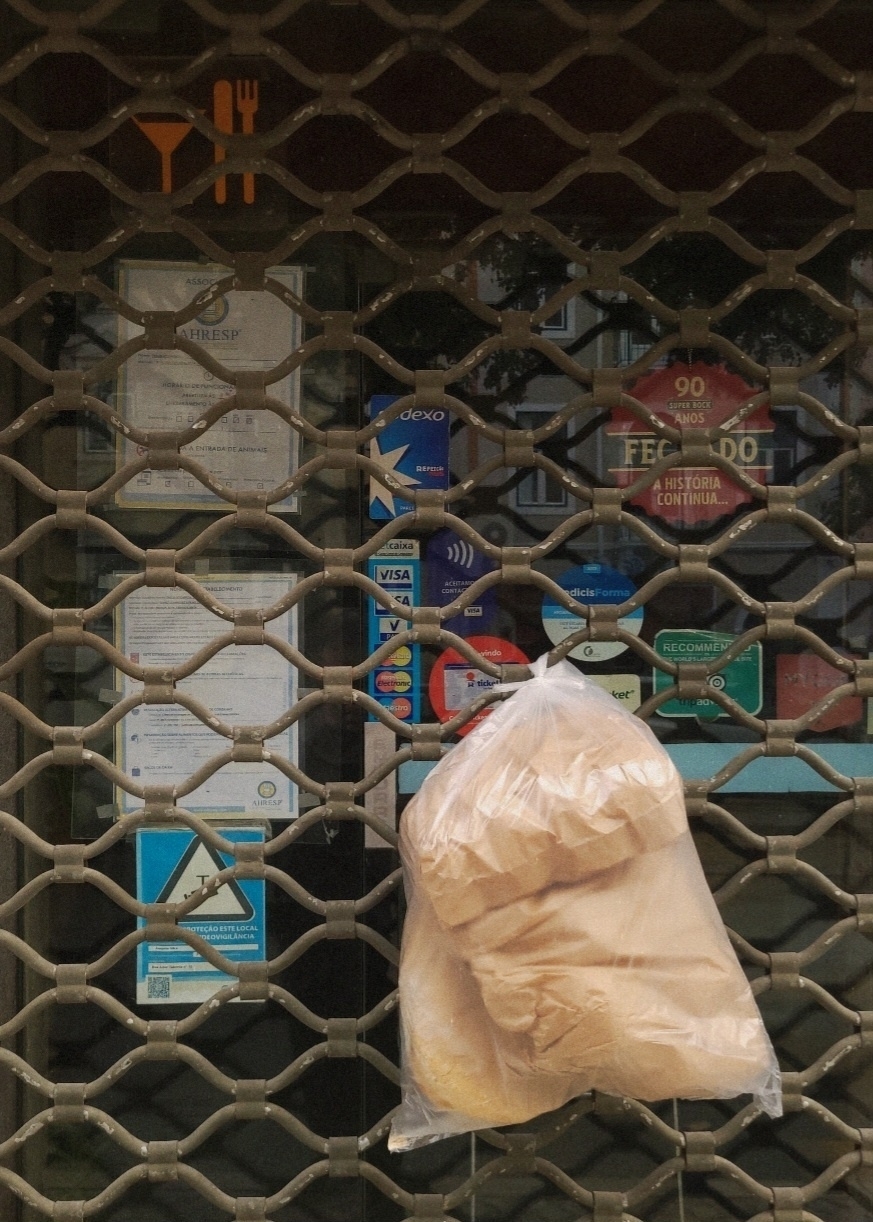 A plastic bag with bread hangs on a metal gate of a restaurant, with various signs and stickers displayed behind it.