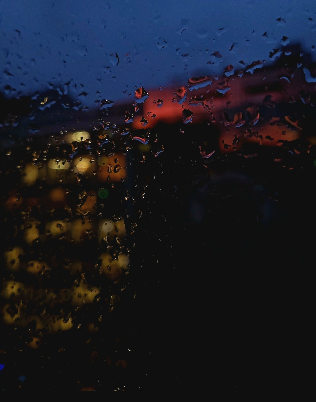 Raindrops on a window blur the view of illuminated buildings against a dark sky at dusk.