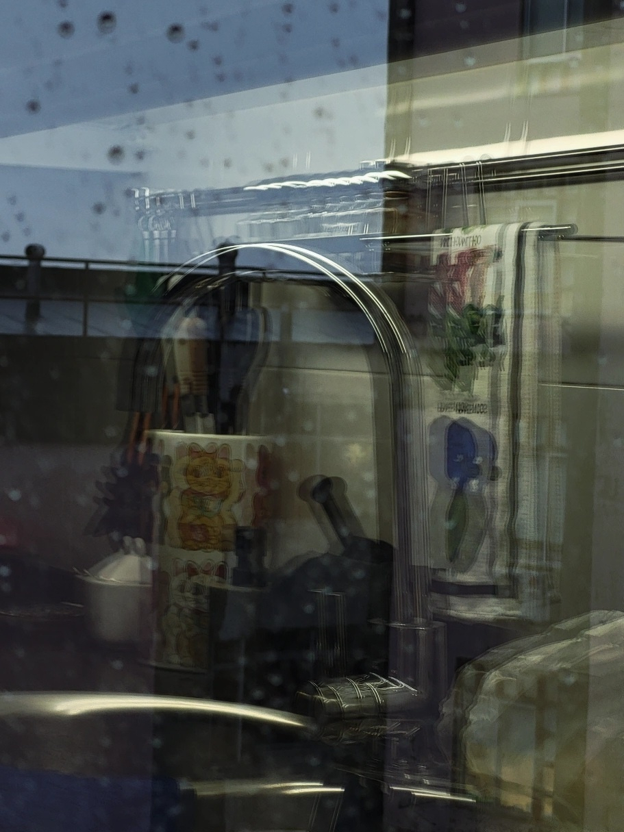 A slightly blurry view of a kitchen sink area with a towel, utensils, and reflections on a wet glass surface.