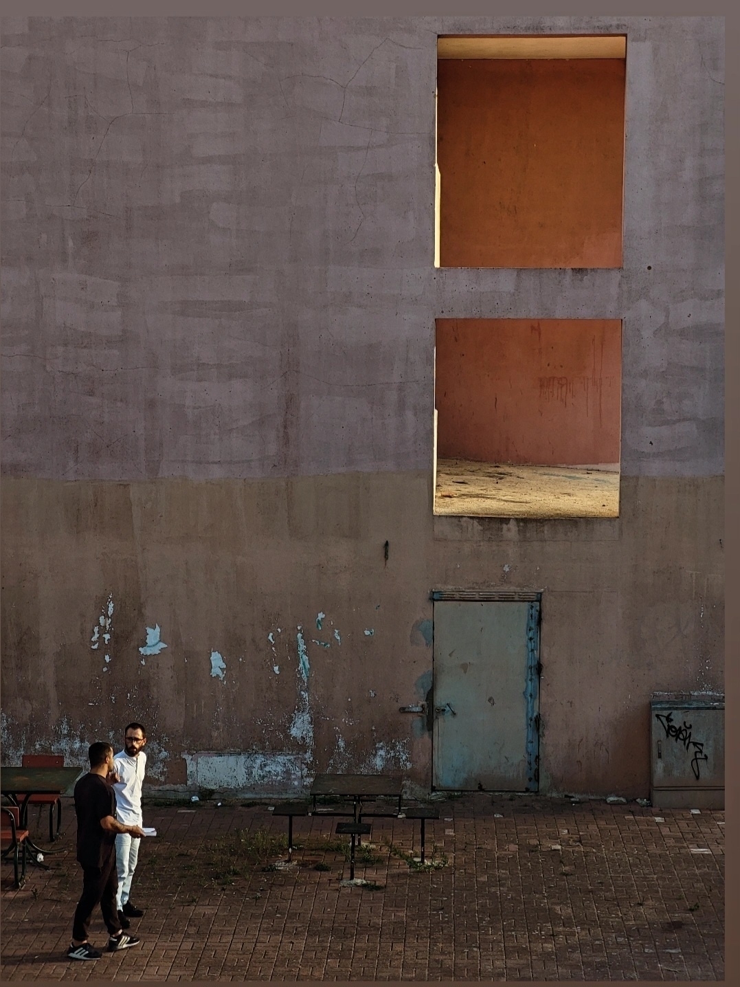 Two people converse in front of a textured wall with high rectangular openings and a weathered door at ground level.