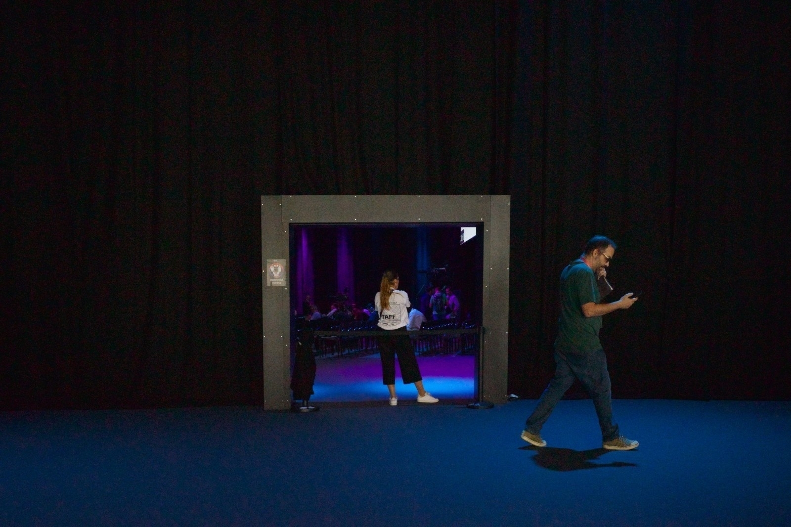A person walks past a doorway where another person stands, with a dimly lit, purple and blue room visible behind them.
