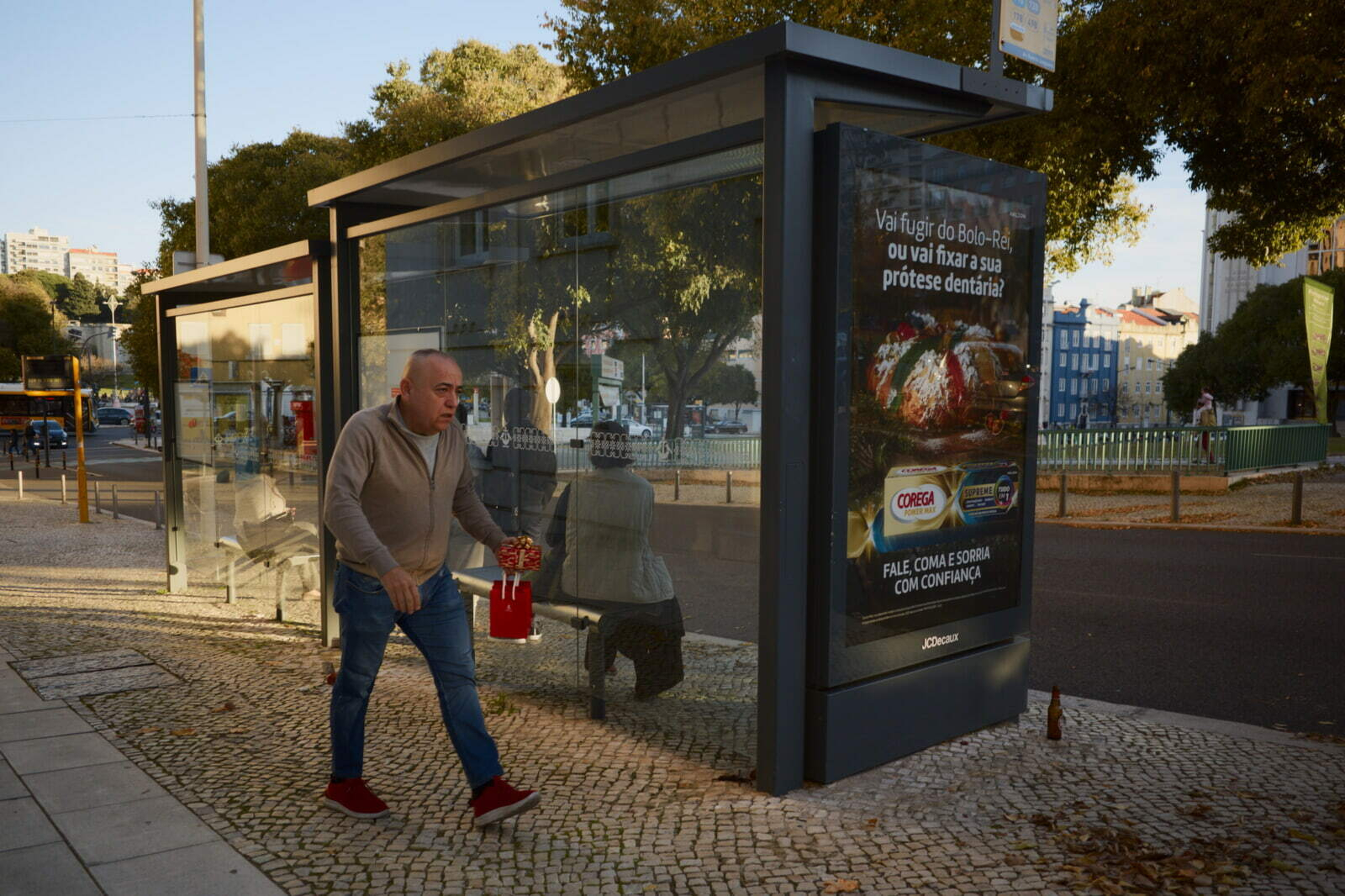A man wearing a beige jacket and blue jeans is walking near a bus shelter with an advertisement for pizza delivery.