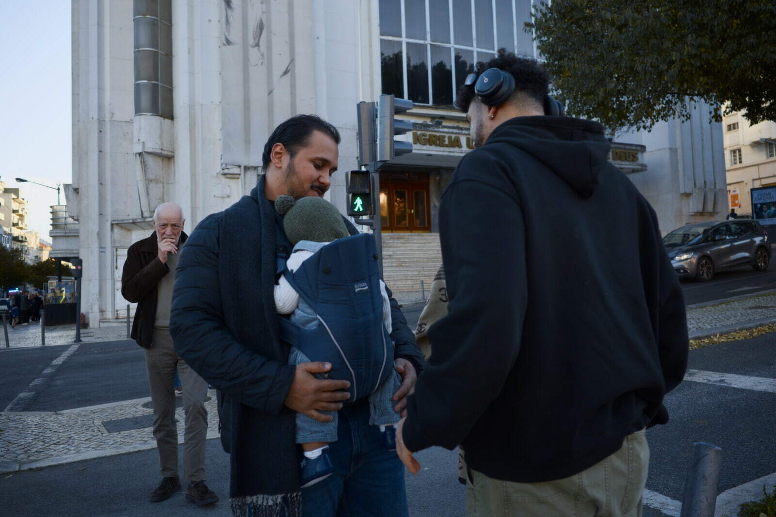 Two people are interacting on a city street, with one holding a baby in a carrier, while another person talks on a phone in the background.