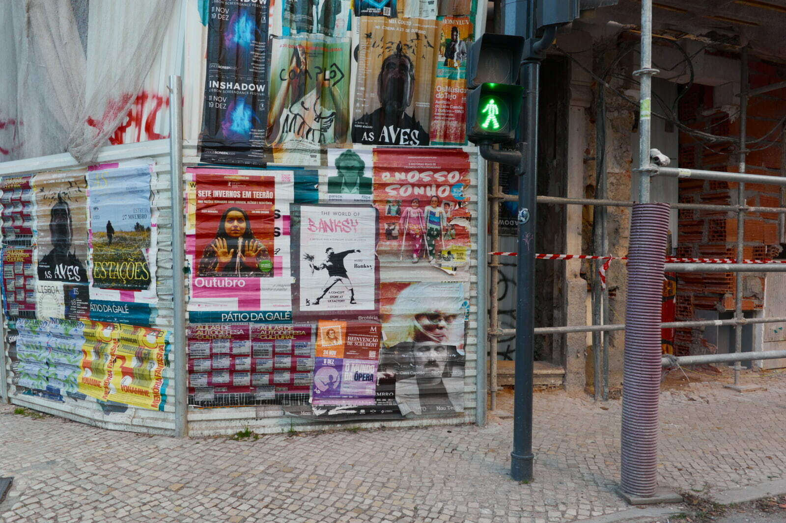 A street corner features a wall covered with colorful posters and a green pedestrian crossing signal beside it.