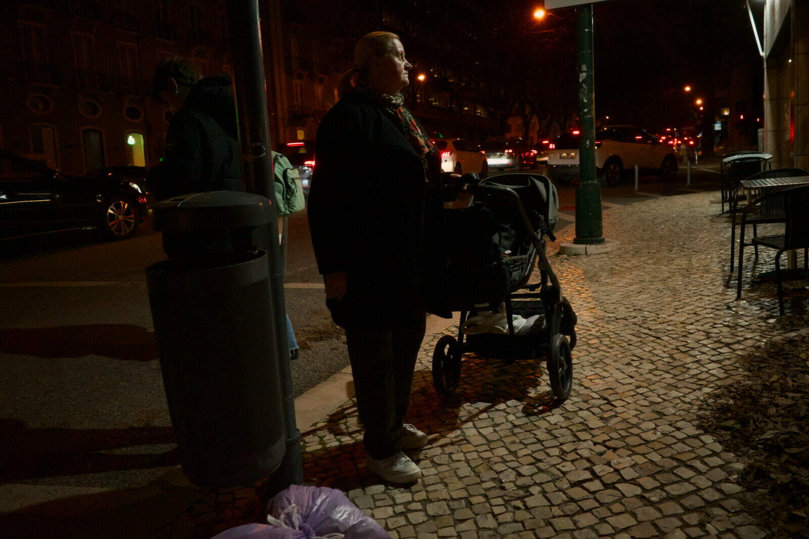 A person stands on a cobblestone sidewalk at night with a stroller near a street with cars.