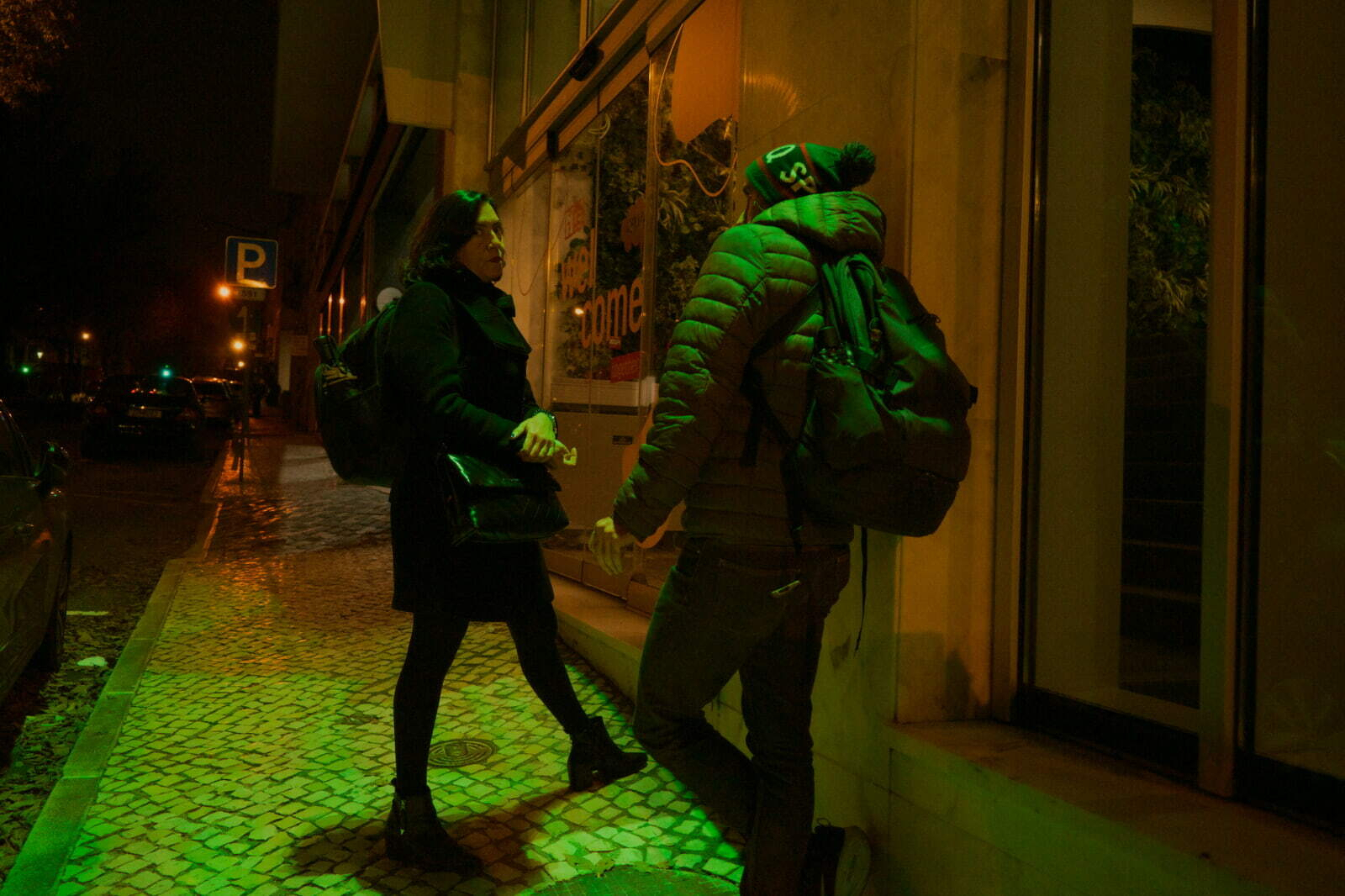 Two people wearing winter clothing are talking on a dimly lit sidewalk at night.