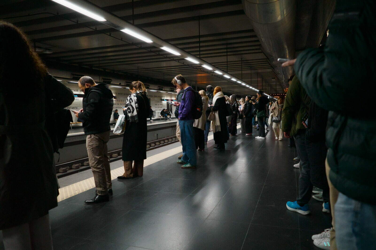 A group of people are waiting on a subway platform, many using their phones.